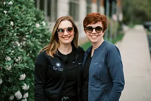 Two women wearing black jackets and sunglasses are standing close to each other on a sidewalk with plants behind them.