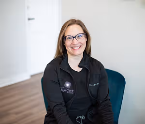A woman with glasses and a black jacket with a logo sits on a blue chair in a room with a white wall and wooden floor.