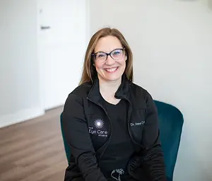 A woman with glasses and a black jacket with a logo sits on a blue chair in a room with a white wall and wooden floor.