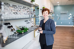 A smiling woman in a blue jacket stands in front of a shelf full of eyeglasses in a store.