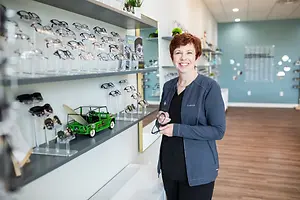 A smiling woman in a blue jacket stands in front of a shelf full of eyeglasses in a store.
