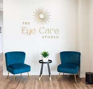 A well-lit room with two blue chairs, a table, and a plant, next to a wall with The Eye Care Studio logo, located on the floor next to a black trash bin.