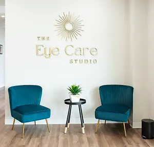 A well-lit room with two blue chairs, a table, and a plant, next to a wall with The Eye Care Studio logo, located on the floor next to a black trash bin.