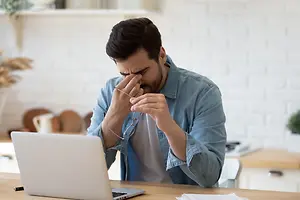 A man sitting at a desk with his hand on his face, holding a pair of glasses