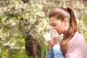 A girl is blowing her nose with a tissue under a tree with white flowers in the background.