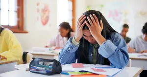 A girl sitting at a desk inside a classroom with her hands covering her face
