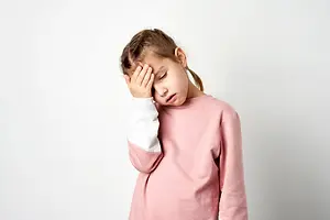 A young girl wearing a pink long-sleeved shirt is standing with her head slightly bowed and her left hand covering her eyes. She seems to be looking at something, maybe a camera. Her hair is tied in a ponytail, and she is standing in a room with white walls.