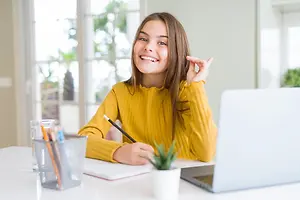 A girl is sitting on a chair in front of a desk and smiling while holding a pencil in her right hand and her left hand is making a hand gesture