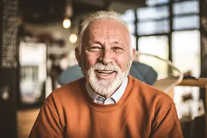 A smiling older man with a white beard and mustache stands in a coffee shop, wearing a warm orange sweater and a white shirt.