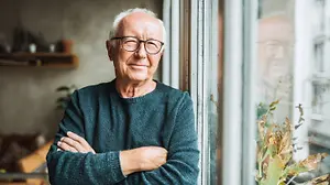 An elderly man wearing glasses and a sweater is smiling and standing in front of a window with his arms crossed.
