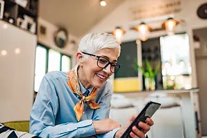 A woman is sitting on a couch and holding a cell phone in her hand while smiling and looking at it.