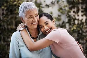 A smiling older woman is hugging a smiling young woman outside, with trees and a white wall in the background.