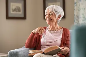 Elderly woman with white hair is sitting on a couch while holding a book and glasses.