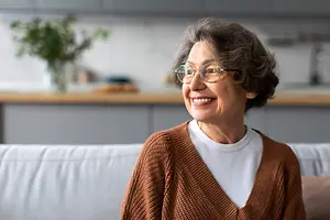 An older woman sitting on a sofa in a living room, smiling with glasses on