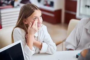 A young girl wearing a white blouse is smiling and touching her face while sitting on a chair in front of a table with a laptop, with another person sitting on the right side of the image.