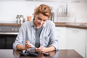 A woman is sitting in the kitchen, holding a blood glucose meter, and looking at it while smiling.