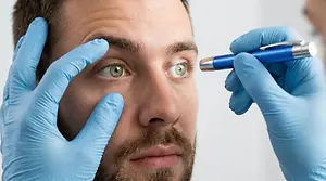 A bearded man wearing blue gloves is undergoing an eye examination by a doctor in a medical setting.