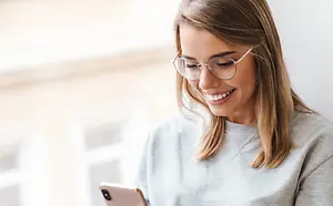 A woman wearing glasses is sitting on a chair and looking at her phone.