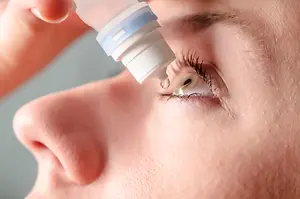 A close-up of a woman's eye with a drop of eye drops being administered by a hand in a room with gray walls