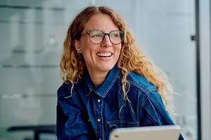 An adult female with curly hair is wearing a denim shirt and glasses, smiling while looking at something on a tablet.