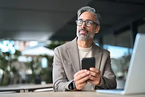 An older man wearing glasses and a gray suit is sitting on a chair, holding a smartphone in his left hand, and looking at it, while his right hand is on his chin. He is probably working at an office.