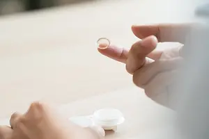 A person wearing contact lenses places a lens on their fingertip, with a contact lens case nearby.