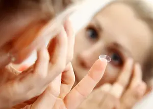A woman is putting a contact lens in her eye while looking in the mirror