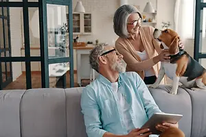 An older couple is sitting on a couch with a beagle dog, the woman is smiling and looking at the man who is looking at a tablet, and they are in a room with a glass door leading to a kitchen area with a cabinet and a table with a potted plant on top.