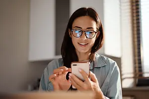 A woman with glasses is smiling while looking at her phone.