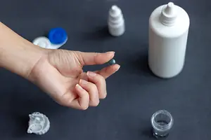 Close up of a hand holding a blue contact lens and a contact lens solution bottle on a table