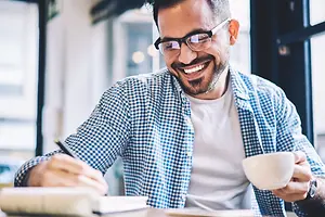 A smiling man in a coffee shop holding a pen and a cup of coffee