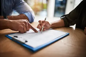 Two people are sitting and pointing at a document on a desk, and one of them is holding a pen and seems to be explaining something.