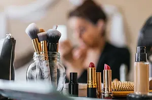 Makeup brushes, lipstick, foundation, and a hairbrush arranged on a vanity with a woman blurred in the background