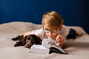 Child wearing glasses reading a book beside a dog on a bed