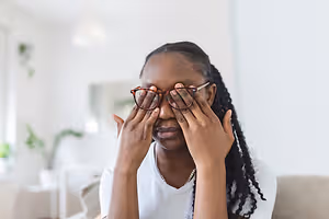 A woman wearing glasses is covering her eyes with her hands while sitting on a couch.
