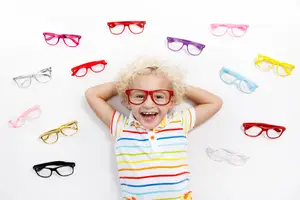 A child lies on a white surface surrounded by a variety of colorful glasses, smiling and wearing red glasses.