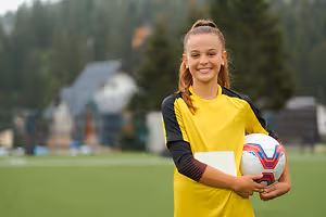 A young girl with a yellow soccer jersey is smiling and holding a soccer ball and a piece of paper on a soccer field with a house and trees in the background.