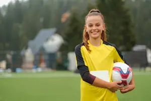 A young girl with a yellow soccer jersey is smiling and holding a soccer ball and a piece of paper on a soccer field with a house and trees in the background.