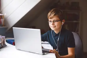 A boy is sitting on a chair in front of a desk with a laptop and wearing earphones