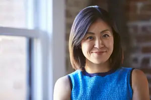 A woman with short black hair is smiling and looking at the camera. She is wearing a blue sleeveless top and is standing in front of a brick wall with a window on the left side.