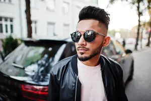 A man wearing sunglasses and a black jacket is standing on the side of the road with a car behind him.