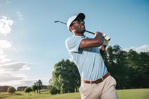 A man in a blue shirt and sunglasses swings a golf club on a golf course.