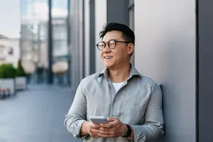 An adult male with glasses and a watch leans against a wall with a cell phone in his hand and smiles
