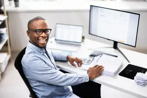 A man sits at a desk in an office with a computer, calculator, and papers. He is smiling and holding a calculator in his right hand. Behind him is a window with a shelf with potted plants on it.