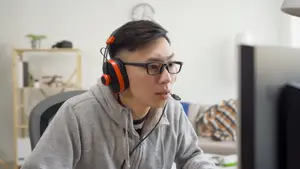 A man in a hoodie is sitting in front of a computer and wearing headphones and glasses.