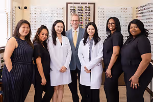 A group of women and a man in white coats and black dresses posing for a photo in a room with glasses on display.