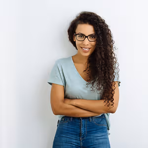Portrait of a smiling woman with curly hair wearing glasses and a blue shirt, standing against a white background.