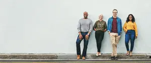 Five people are standing in a line in front of a white wall. The man on the left is wearing a watch and sneakers, the woman in the middle is wearing a yellow shirt, the man in the middle is wearing a denim jacket, the man on the right is wearing a brown shirt, and the woman on the right is wearing blue pants.