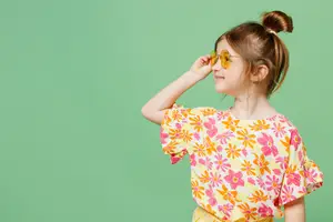 A young girl with a bun hairstyle wearing a yellow floral dress and yellow sunglasses is smiling and posing in front of a green wall