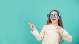 A girl with brown hair wearing a white long-sleeve shirt and blue heart-shaped glasses is pointing with her left hand in front of a green background. She has her mouth open, probably shouting at someone.